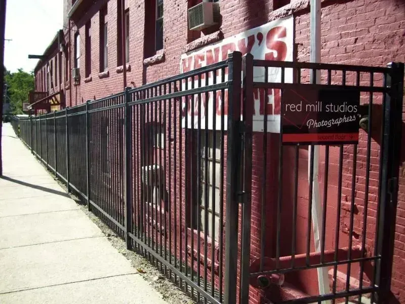 A black metal fence runs along a sidewalk beside a red brick building with a