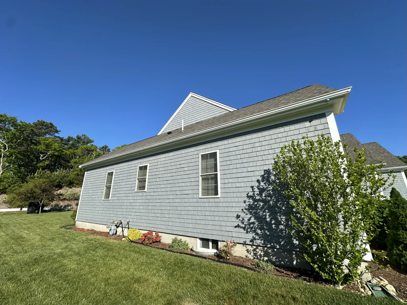 Side view of a gray house with vinyl siding and multiple windows, surrounded by a well-maintained lawn, a bush, and a garden bed with flowers, under a clear blue sky.
