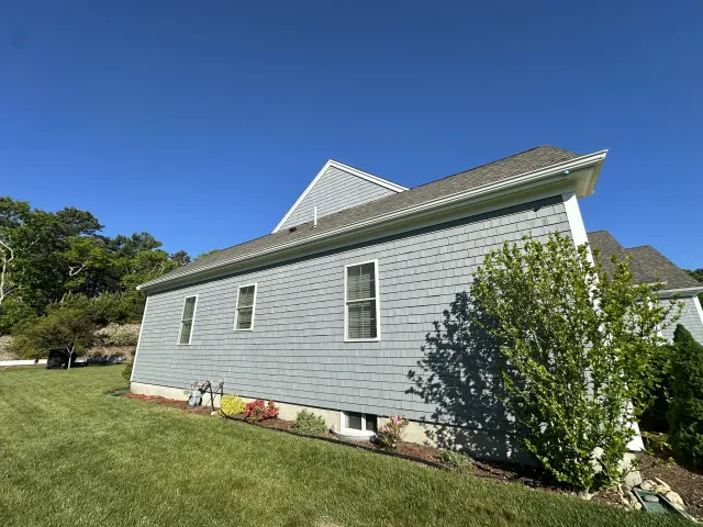 Side view of a gray house with vinyl siding and multiple windows, surrounded by a well-maintained lawn, a bush, and a garden bed with flowers, under a clear blue sky.