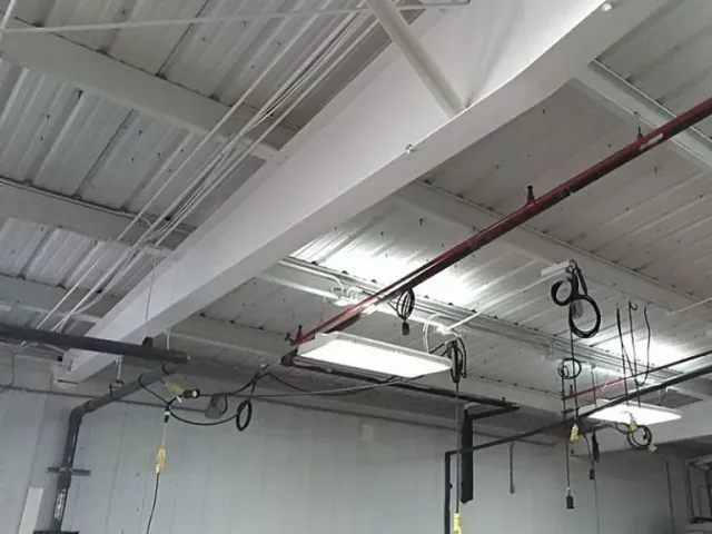 Interior view of a warehouse ceiling with metal beams, pipes, wiring, and fluorescent lights. The space is mostly white and gray.