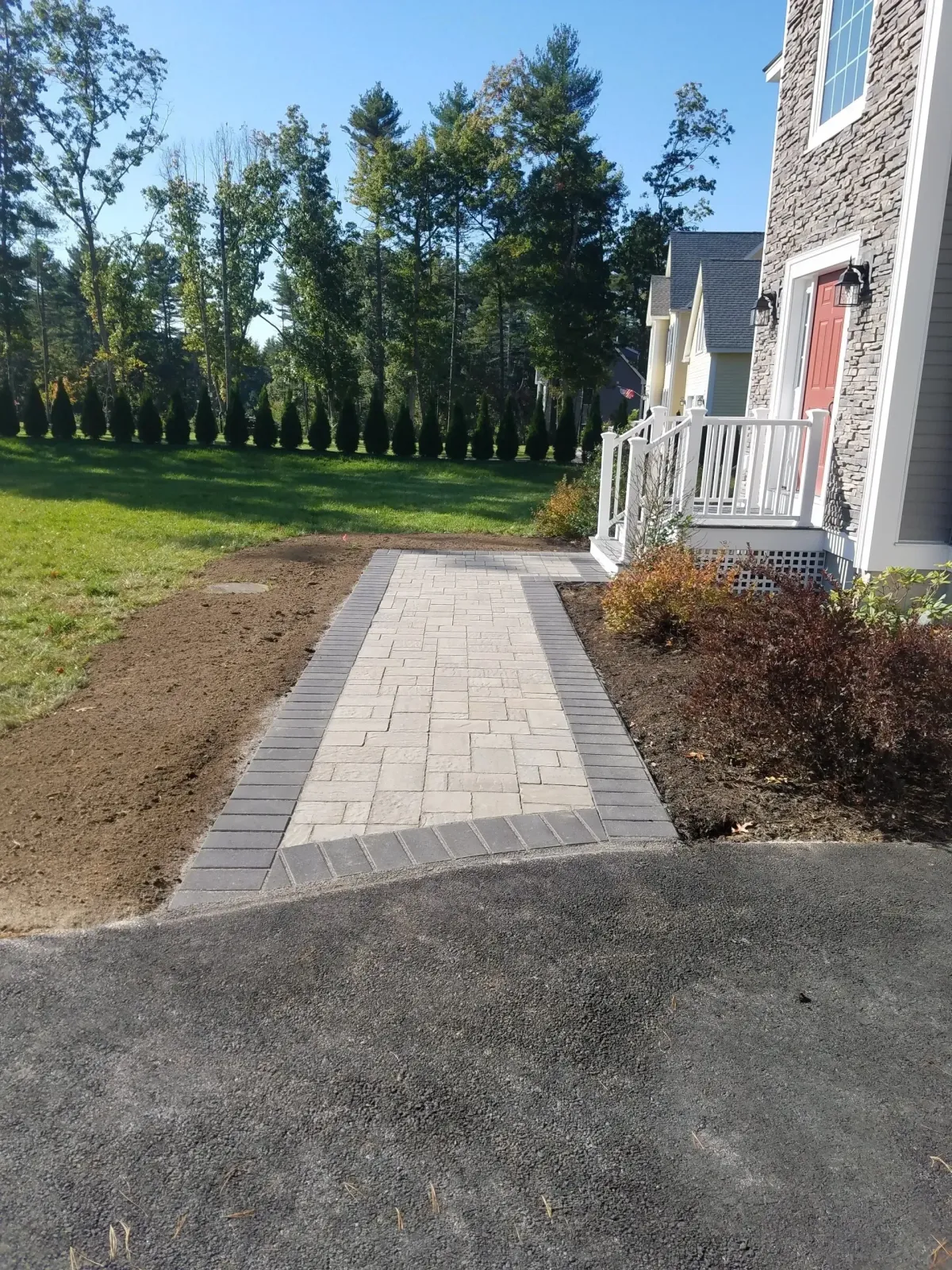 Paver walkway leading to a house entrance, bordered by grass, shrubs, and gravel driveway.