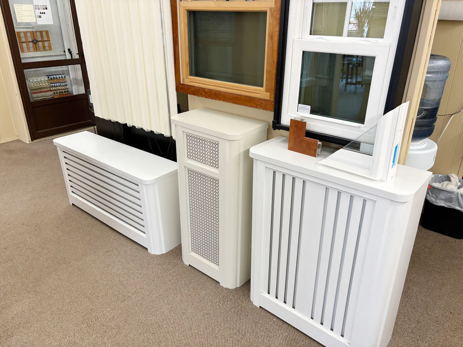 Three white radiator covers in different styles displayed in the showroom below sample windows