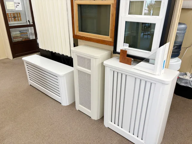 Three white radiator covers in different styles displayed in the showroom below sample windows