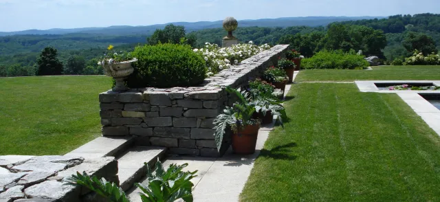 Beautiful garden with stone steps, potted plants, a stone wall, green grass lawn, and scenic mountains in the background.