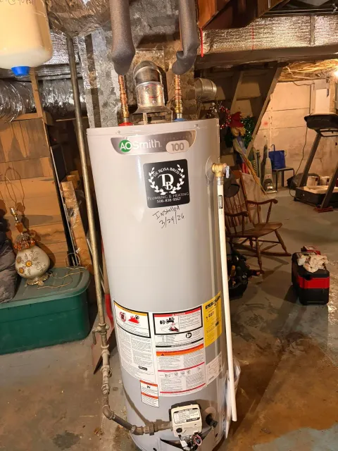 A newly installed gas water heater standing in a basement with its flue elbow, copper supply connections and temperature and pressure relief pipe fitted, and a De Rosa Bros sticker on the front of the tank.