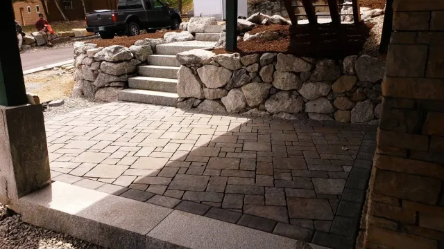 A stone patio features two patterns of pavers leading to stone steps and a retaining wall in a partially shaded outdoor area.