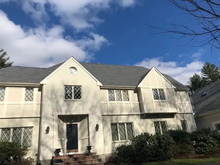 Two-story beige house with dark roof, white trim, and front entrance under a blue sky