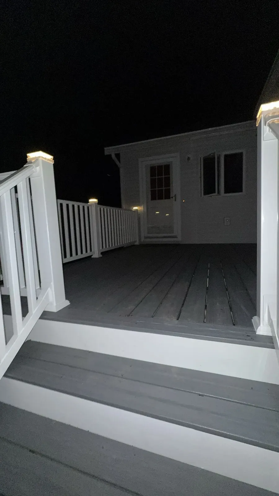 A wooden deck with white railings and stairs at night. A house with a door and windows is visible in the background, illuminated by outdoor lights.