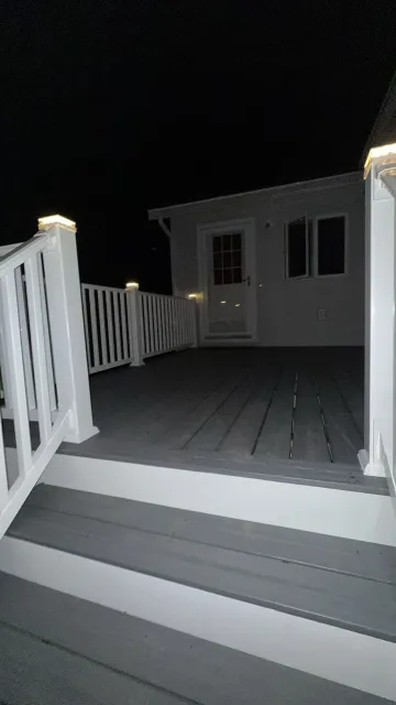 A wooden deck with white railings and stairs at night. A house with a door and windows is visible in the background, illuminated by outdoor lights.