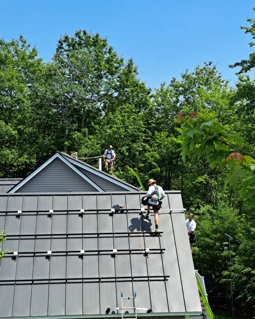 Workers installing solar panel mounts on a rooftop surrounded by trees.