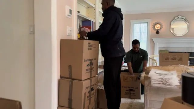 Two movers pack items in a living room, one arranging books on a shelf, the other placing items into a box, with moving boxes and household furniture around.