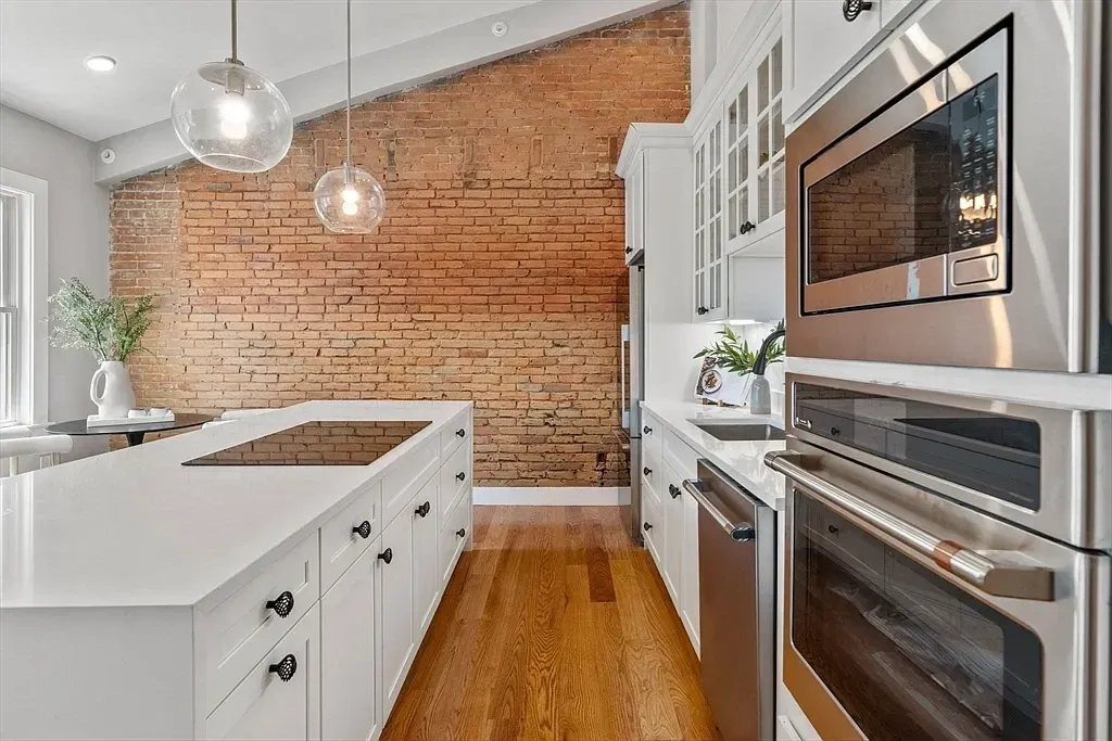 Kitchen with white cabinets, stainless steel appliances, a brick accent wall, wooden floor, and modern glass pendant lights.