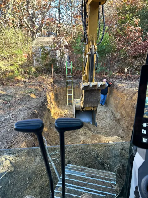 An excavator digging a trench; a person stands inside. Yellow machinery, brown soil, and a green ladder.