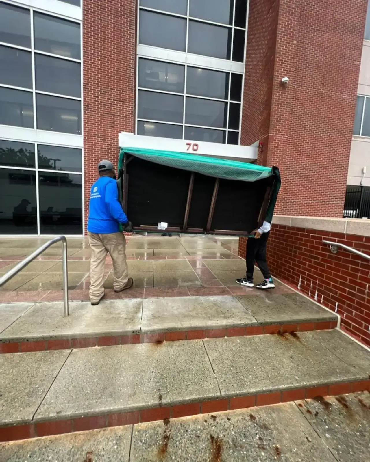 Two people carrying a large, wrapped object up concrete steps in front of a building with a number 70.