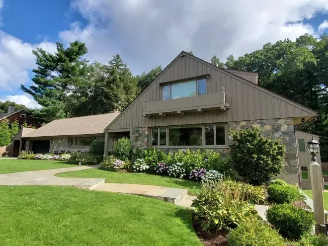 House with stone exterior and tan roof, surrounded by greenery under a cloudy sky.