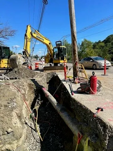 Construction site with an excavator digging a trench, orange traffic cones, a fire extinguisher, and utility pole during daytime.