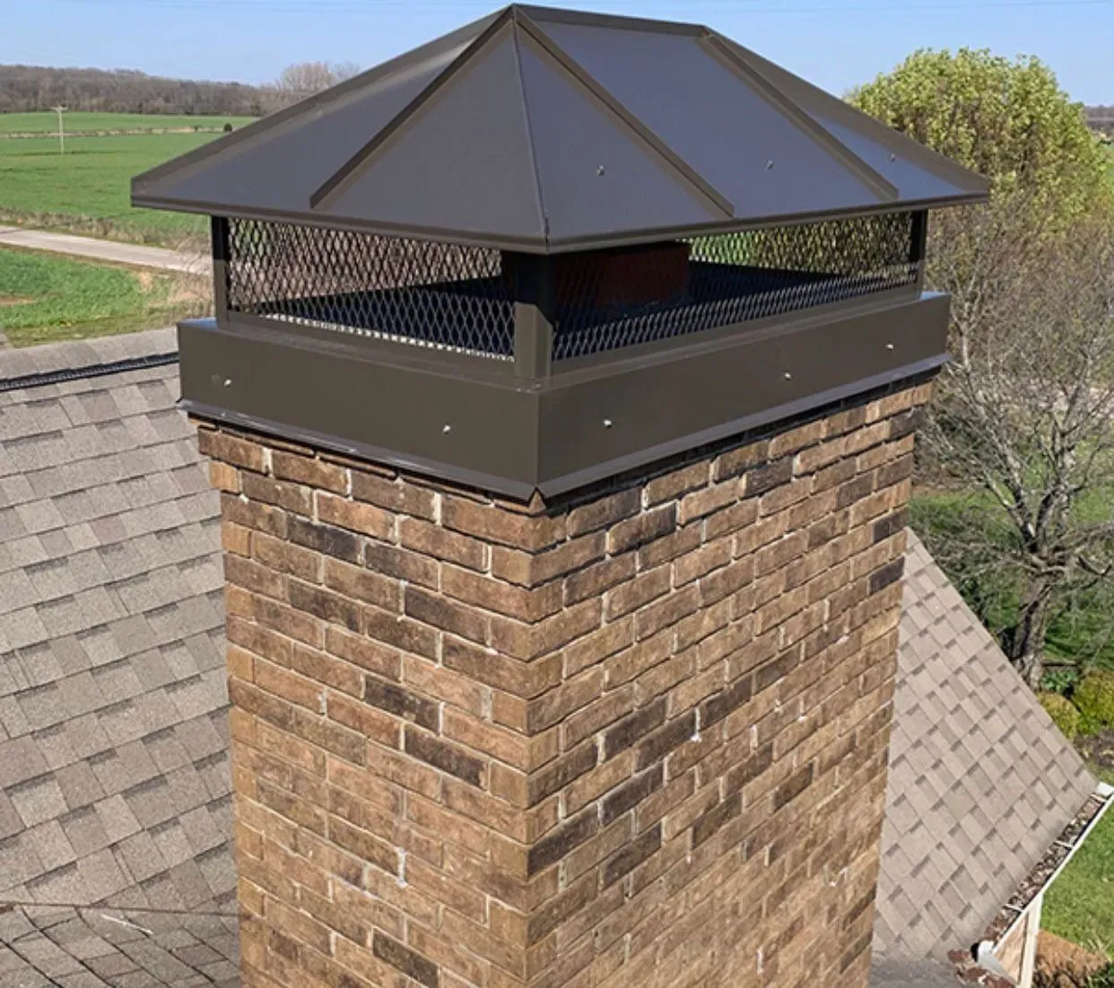 Brick chimney with dark metal cap on a shingled roof, set against a rural landscape