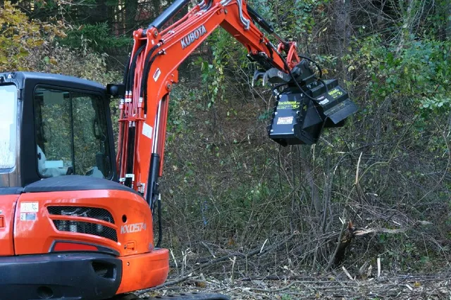 A red Kubota KX057-4 excavator with a vegetation mulching attachment is working near dense shrubs and trees.