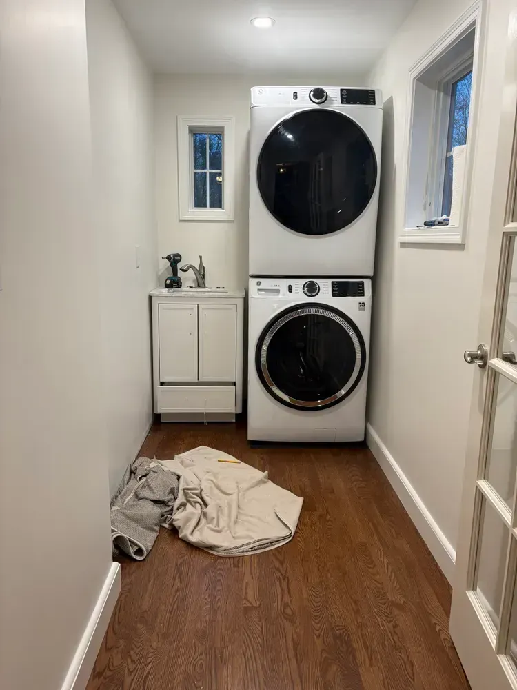 A narrow laundry room with a stacked washer and dryer, a small white vanity sink, and clothing on the wood floor.