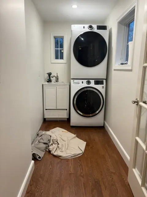 A narrow laundry room with a stacked washer and dryer, a small white vanity sink, and clothing on the wood floor.
