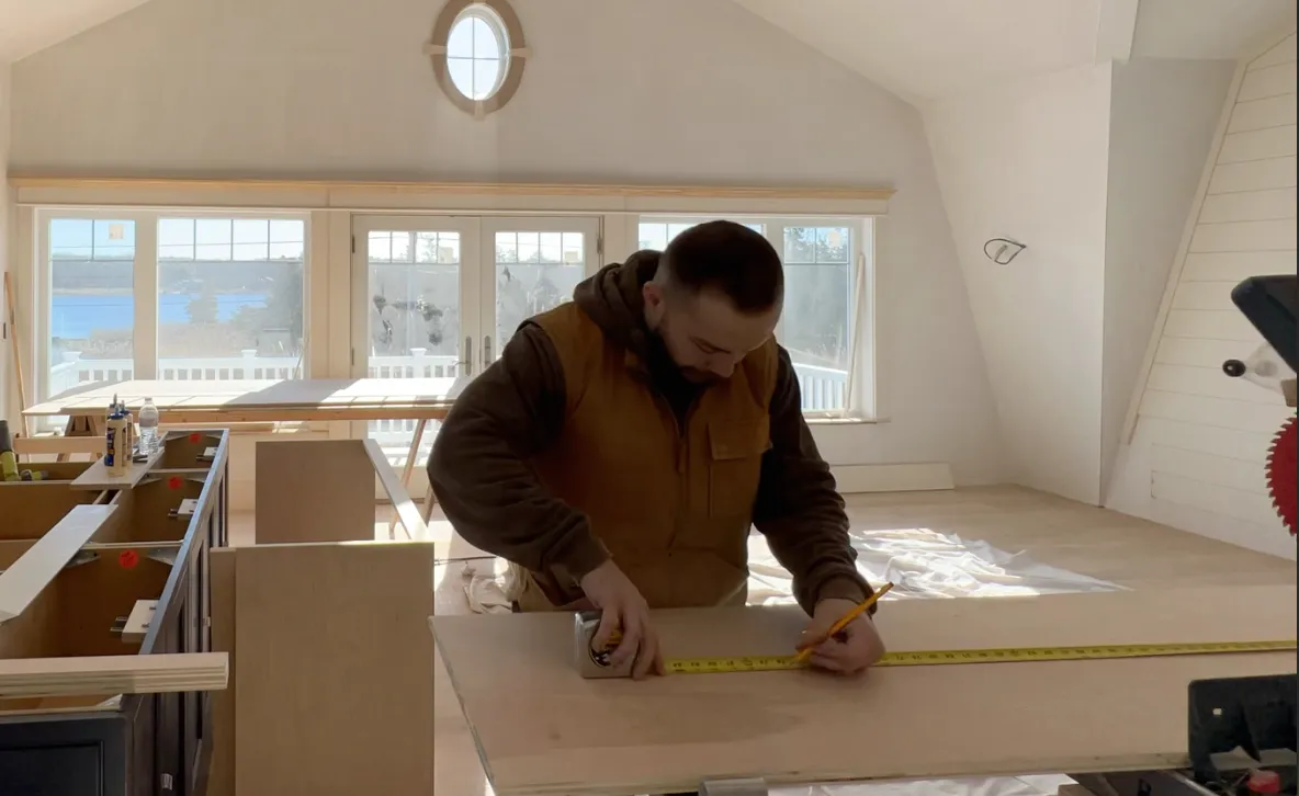 Man measuring a wooden board with a yellow tape measure and holding a pencil in a bright room with large windows.
