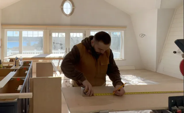 Man measuring a wooden board with a yellow tape measure and holding a pencil in a bright room with large windows.