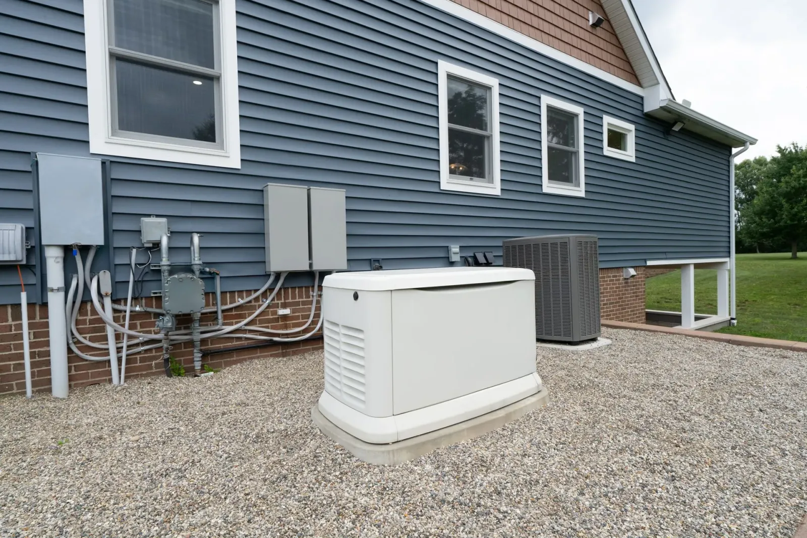 White backup generator on gravel beside blue house with utility meters and siding.