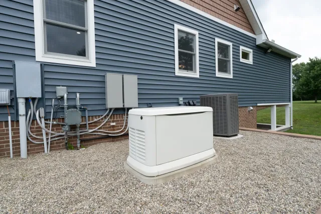 White backup generator on gravel beside blue house with utility meters and siding.