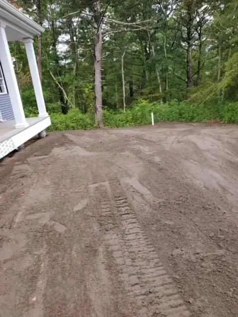 Freshly graded dirt driveway beside a house, with tire tracks and trees in the background.