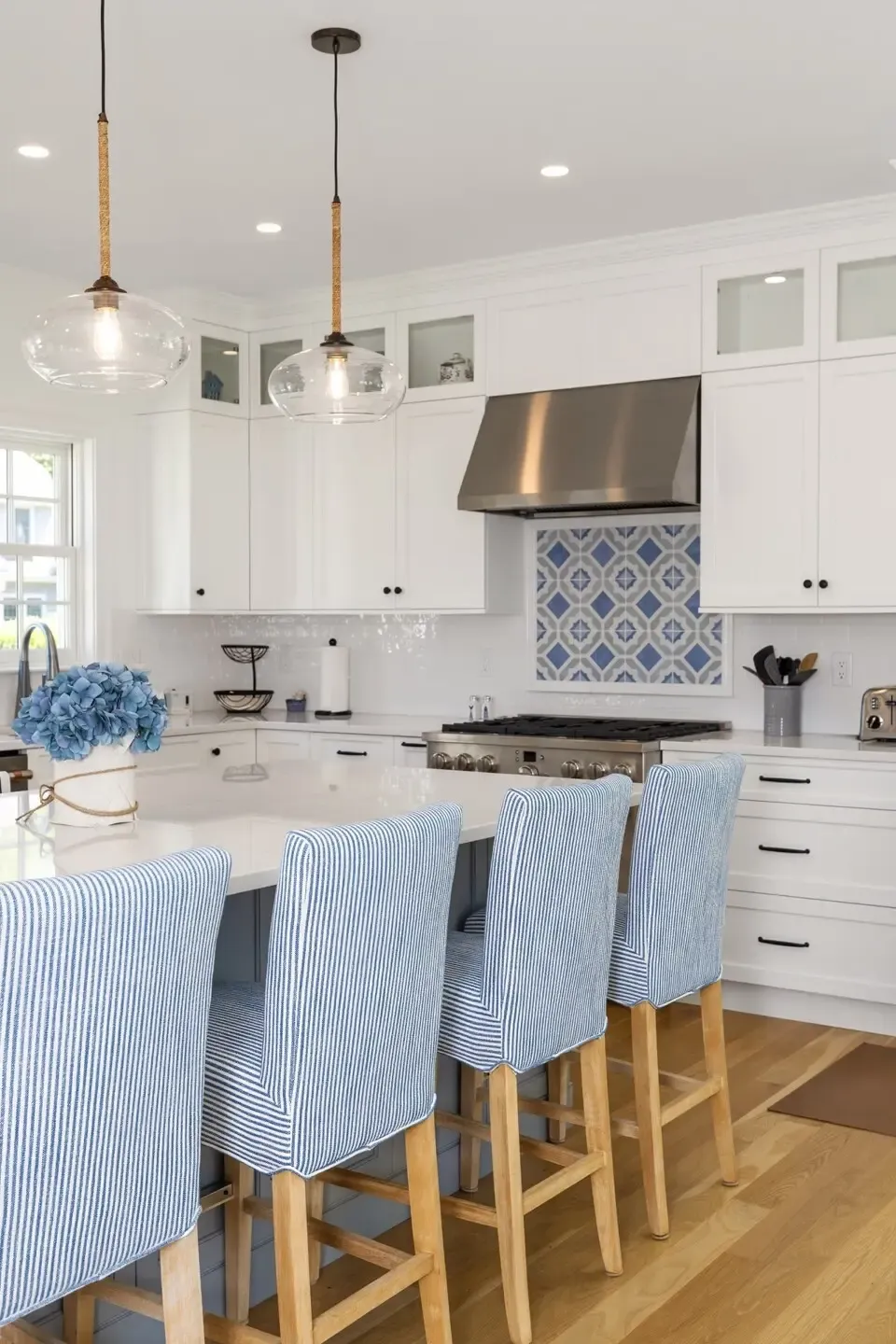 Bright white kitchen with a blue tiled backsplash, stainless range, and island seating; light blue striped bar stools and glass pendant lights.