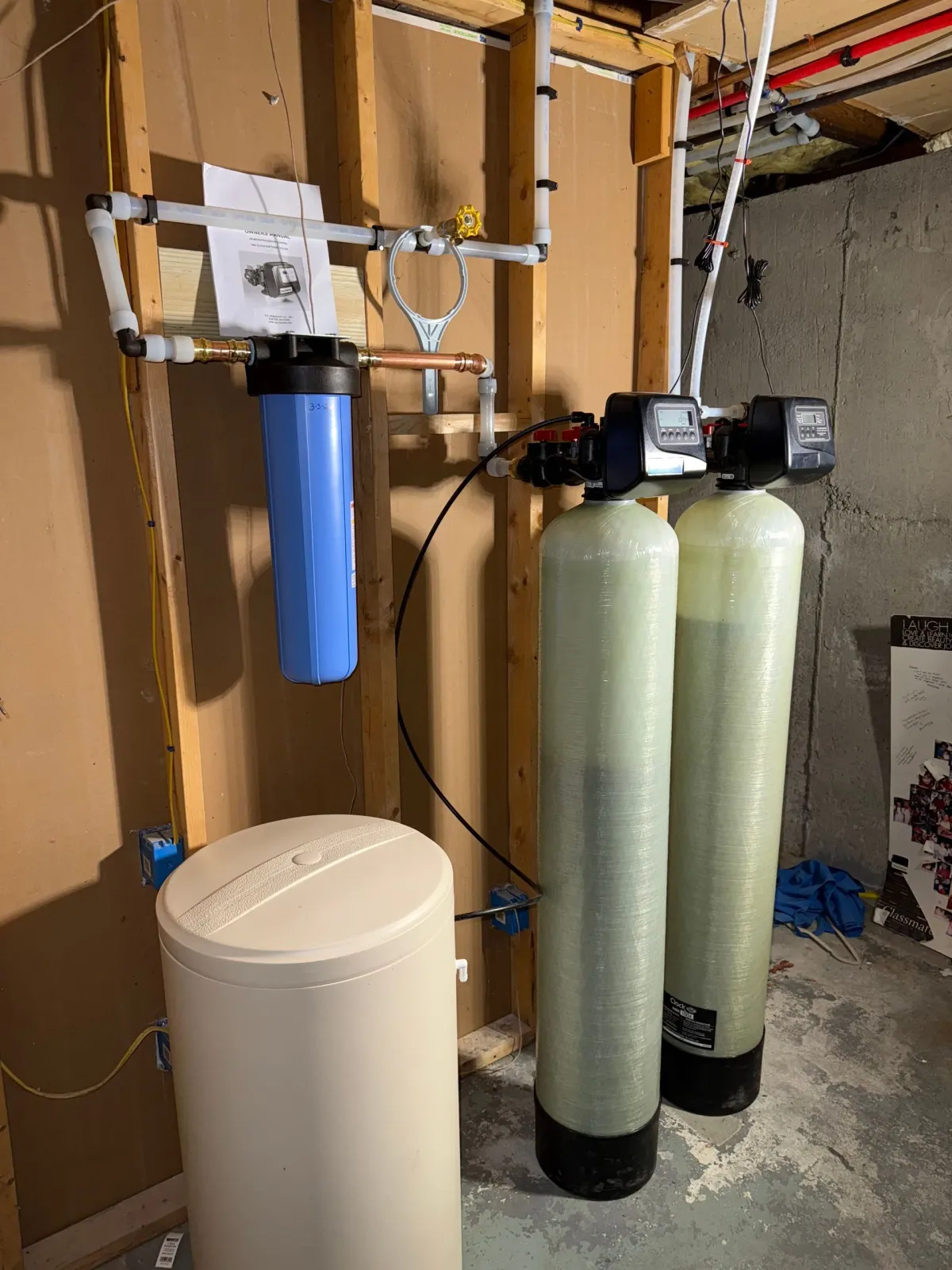 A basement water filtration system featuring a blue pre-filter, two tall treatment tanks, and a beige brine tank.