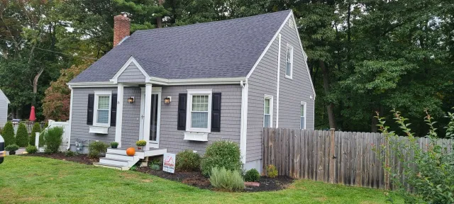 Gray house with a dark roof and a picket fence in front of greenery.