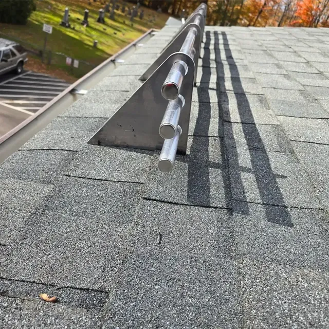 A metal snow guard with three horizontal rods mounted on a gray shingled roof near a gutter.