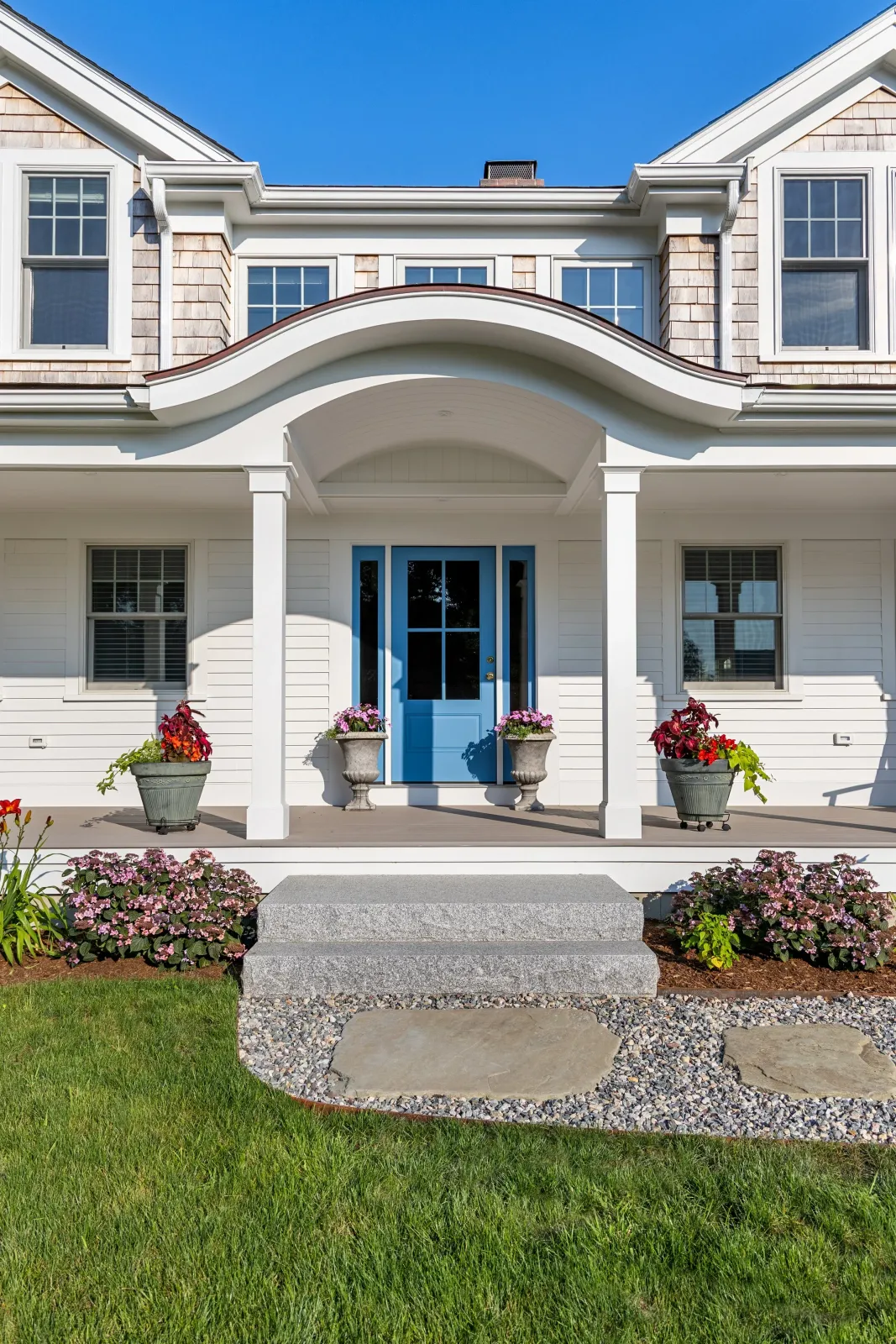 Front porch of a house with a bright blue door, white siding, and a curved roof. Flower pots with colorful flowers are on either side of the door, and there are flower beds with pink and red flowers in the front yard. Stone steps lead up to the porch, and the sky is clear blue.
