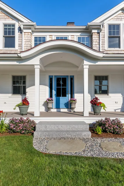 Front porch of a house with a bright blue door, white siding, and a curved roof. Flower pots with colorful flowers are on either side of the door, and there are flower beds with pink and red flowers in the front yard. Stone steps lead up to the porch, and the sky is clear blue.