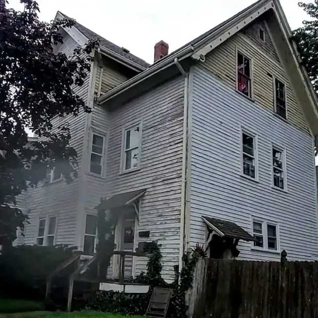 Severely weathered three-story clapboard home with peeling paint, damaged gutters, and deteriorating wood siding in need of exterior painting in Rhode Island