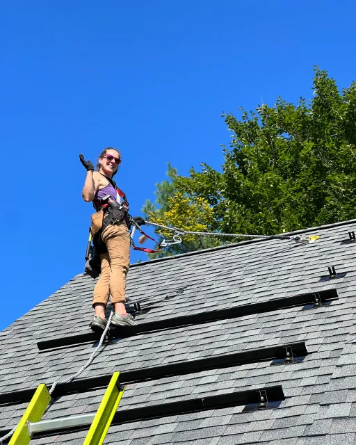A woman standing on a roof while working on solar panel mounts, wearing safety gloves, harness, and sunglasses, with trees and a clear blue sky in the background.