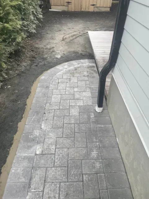A new grey stone paver walkway runs alongside a house exterior wall and a wooden deck, with unlandscaped dirt to the left.