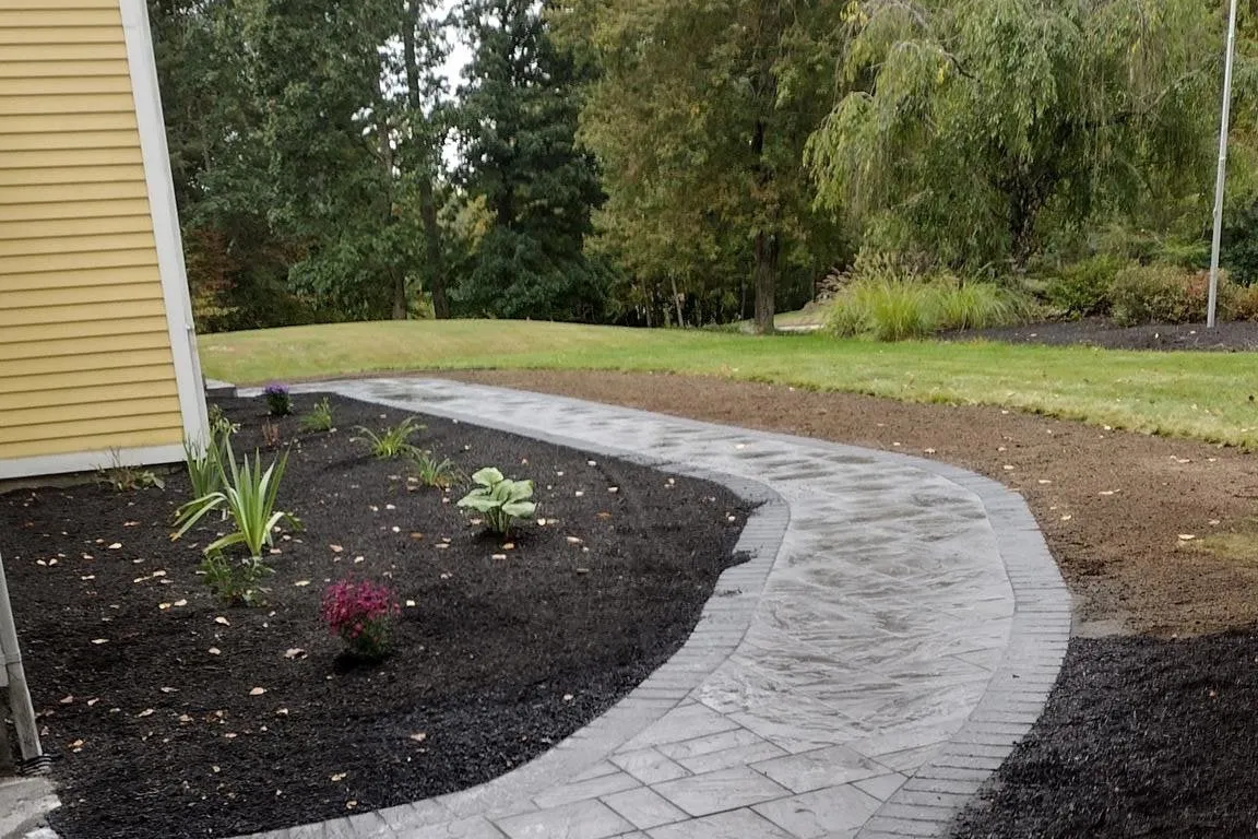 S-curved stamped concrete walkway connecting a dark asphalt driveway to a yellow-sided house in Charlton, MA, surrounded by fresh black mulch, small plants, green grass, and a backdrop of dense trees.