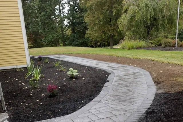 S-curved stamped concrete walkway connecting a dark asphalt driveway to a yellow-sided house in Charlton, MA, surrounded by fresh black mulch, small plants, green grass, and a backdrop of dense trees.