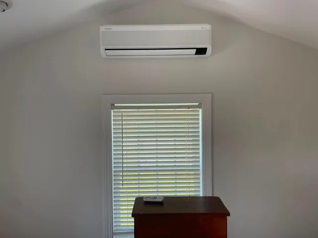 Small room with a wall-mounted air conditioner above a window with blinds and a wooden dresser below.