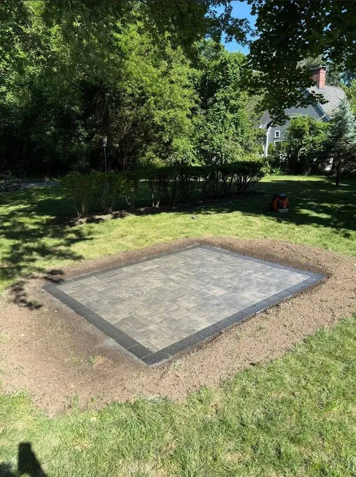 Small concrete slab bordered by brick in a grassy backyard with trees and a house in the background