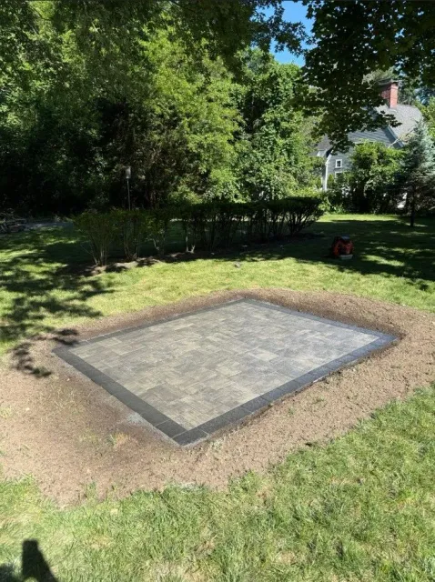 Small concrete slab bordered by brick in a grassy backyard with trees and a house in the background