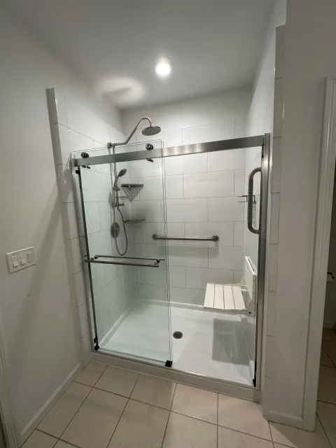 Clean walk-in shower with glass door, white tiled walls, overhead showerhead, handheld shower, a corner shelf, and a built-in bench inside a bathroom.