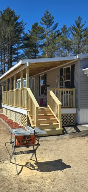 Mobile home with wood deck, railings, and covered porch under clear blue sky with pine trees in background