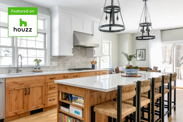 A bright modern kitchen with white upper cabinets, wood lower cabinets, a white island with bookshelves, and a seating area with woven chairs around the island. Two black industrial pendant lights hang from the ceiling. Large windows allow natural light and a view of a snowy outdoor landscape.