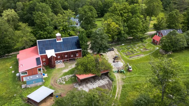 Aerial view of a farm with a large red barn equipped with solar panels, a smaller red shed, a vegetable garden, a horse riding area, and multiple trees surrounding the farm on a sunny day.