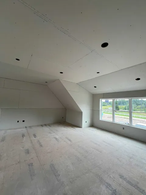 Interior view of a room under construction with drywall installed.  Unfinished walls, ceiling with recessed light openings, and a window.