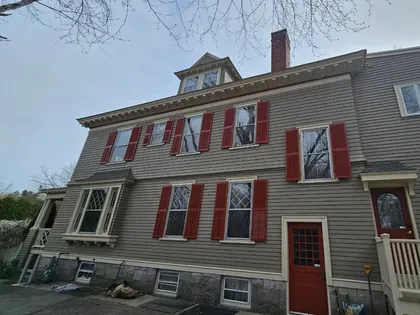 Gray house with red shutters and a red door, viewed from the front under a cloudy sky