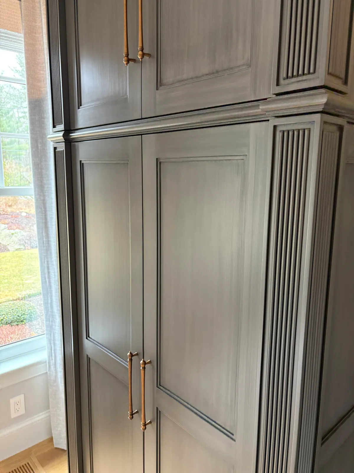 A tall built-in wooden cabinet with paneled doors and vertical grooves, featuring brass handles, situated next to a window with sheer curtains.